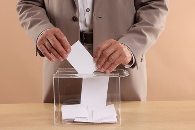 Referendum. Man putting his vote into ballot box at wooden table against beige background, closeup