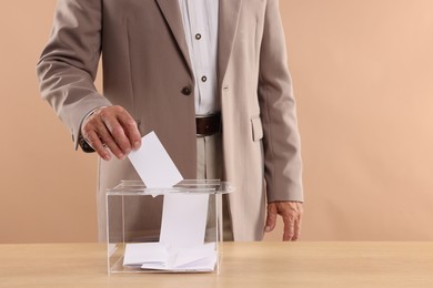 Photo of Referendum. Man putting his vote into ballot box at wooden table against beige background, closeup