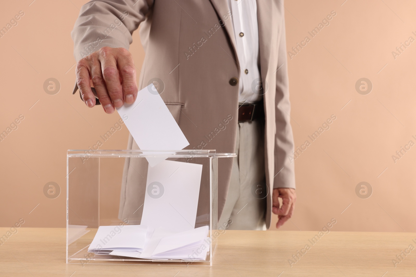 Photo of Referendum. Man putting his vote into ballot box at wooden table against beige background, closeup