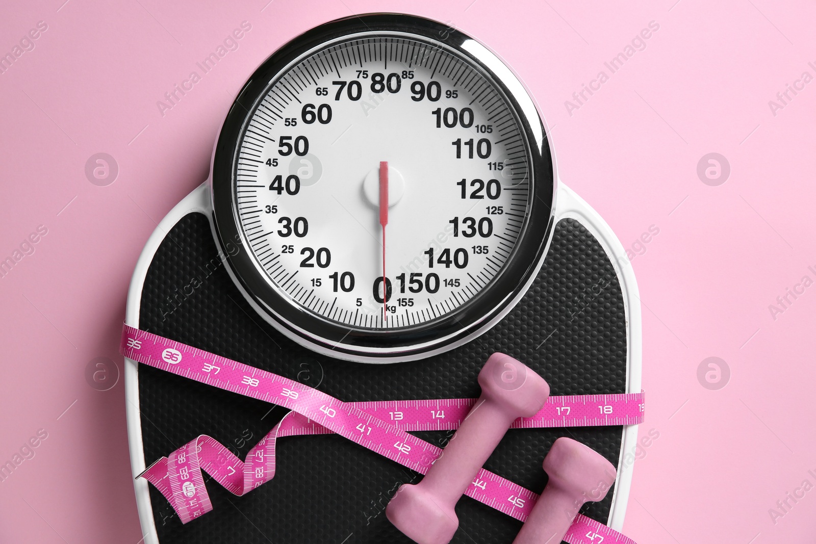 Photo of Scale tied with measuring tape and dumbbells on pink background, top view