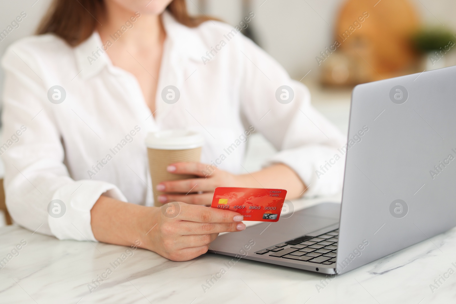 Photo of Online banking. Woman with credit card, paper cup and laptop paying purchase at white marble table, closeup