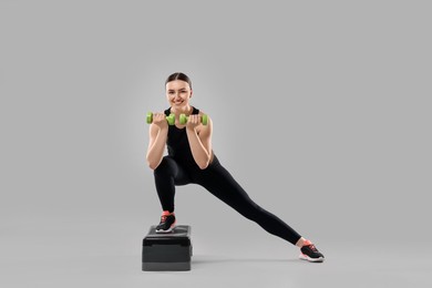 Photo of Young woman doing aerobic exercise with dumbbells and step platform on light background