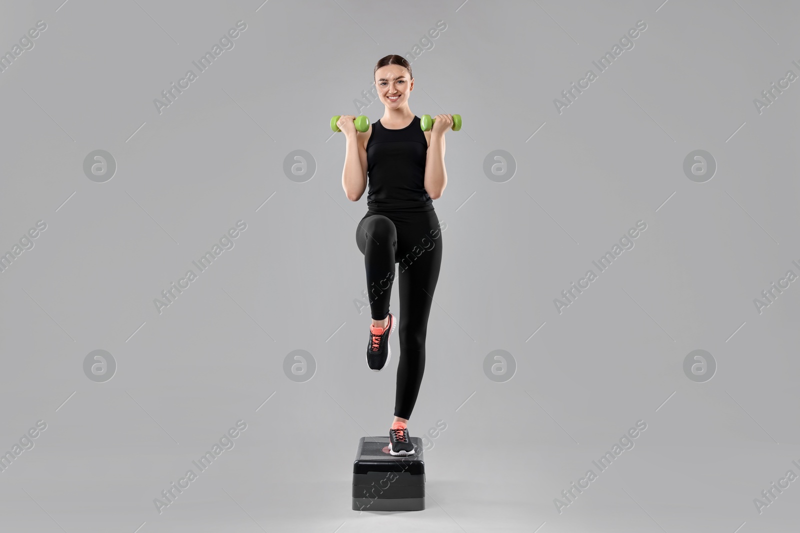 Photo of Young woman doing aerobic exercise with dumbbells and step platform on light background