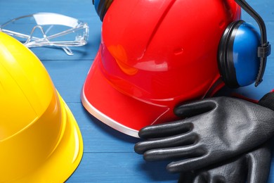 Photo of Hard hats, protective gloves, earmuffs and goggles on blue wooden background, closeup
