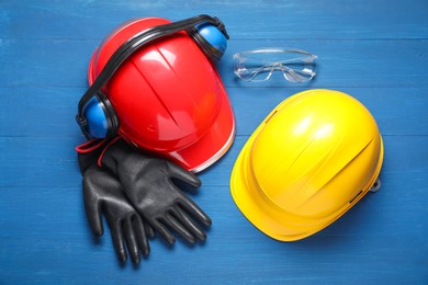 Hard hats, protective gloves, earmuffs and goggles on blue wooden background, flat lay