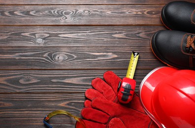 Pair of working boots, hard hat, protective gloves, goggles and tape measure on wooden background, flat lay. Space for text