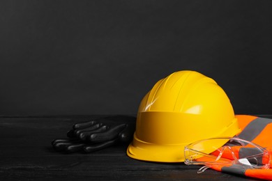 Reflective vest, hard hat, protective gloves and goggles on black wooden surface against gray background, space for text