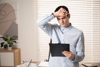 Photo of Embarrassed man with clipboard and pen in office, space for text