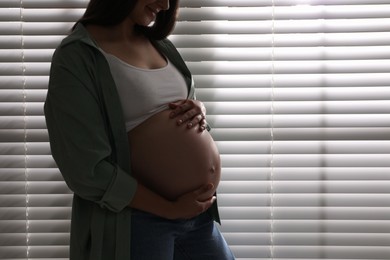 Photo of Beautiful pregnant woman near window blinds at home, closeup. Space for text