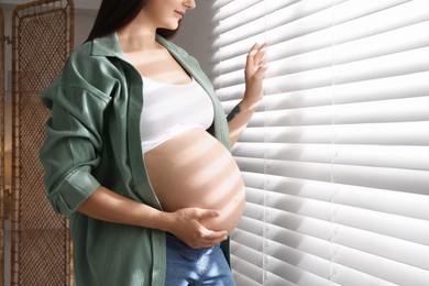 Pregnant woman near window blinds at home, closeup