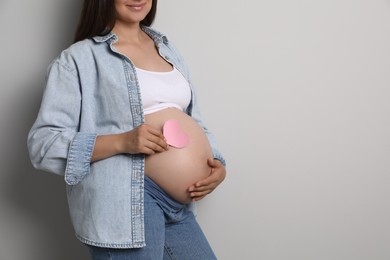 Photo of Pregnant woman with pink paper heart on gray background, closeup. Space for text