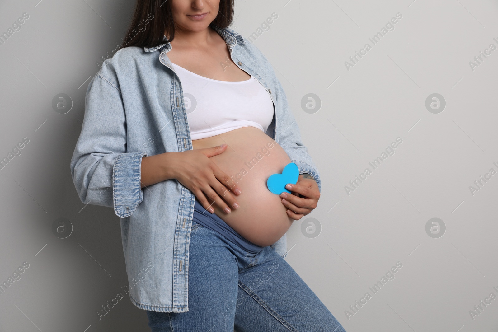 Photo of Pregnant woman with light blue paper heart on gray background, closeup. Space for text