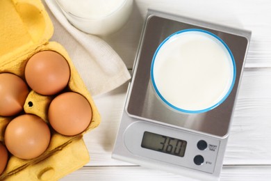 Kitchen scale with glass of milk near eggs on white wooden table, flat lay