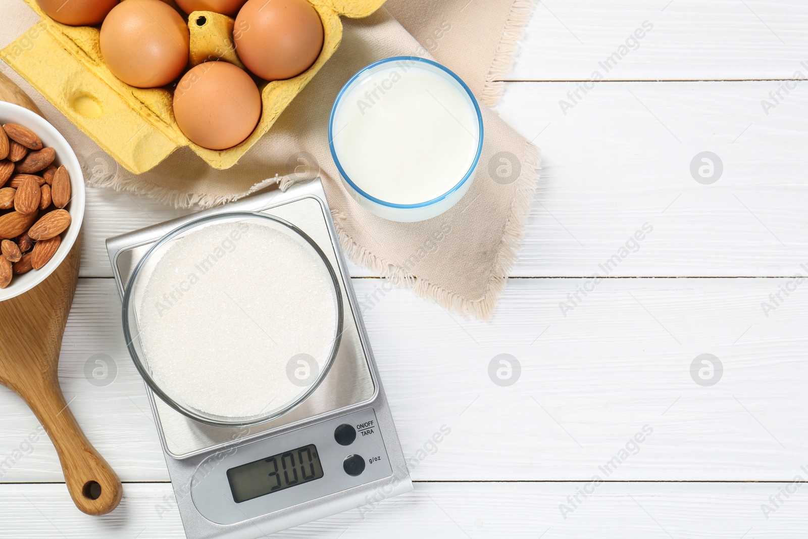 Photo of Kitchen scale with bowl of sugar among milk, almond and eggs on white wooden table, flat lay. Space for text