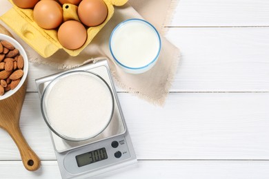 Kitchen scale with bowl of sugar among milk, almond and eggs on white wooden table, flat lay. Space for text