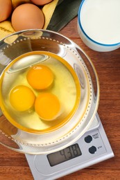 Photo of Kitchen scale with bowl of raw eggs and milk on wooden table, flat lay