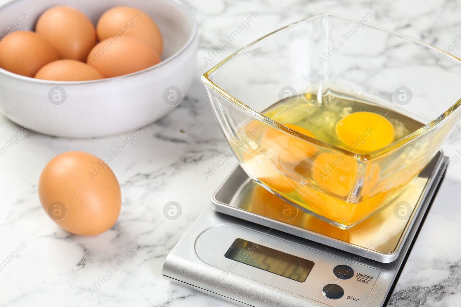 Photo of Kitchen scale with bowl of raw eggs on white marble table, closeup