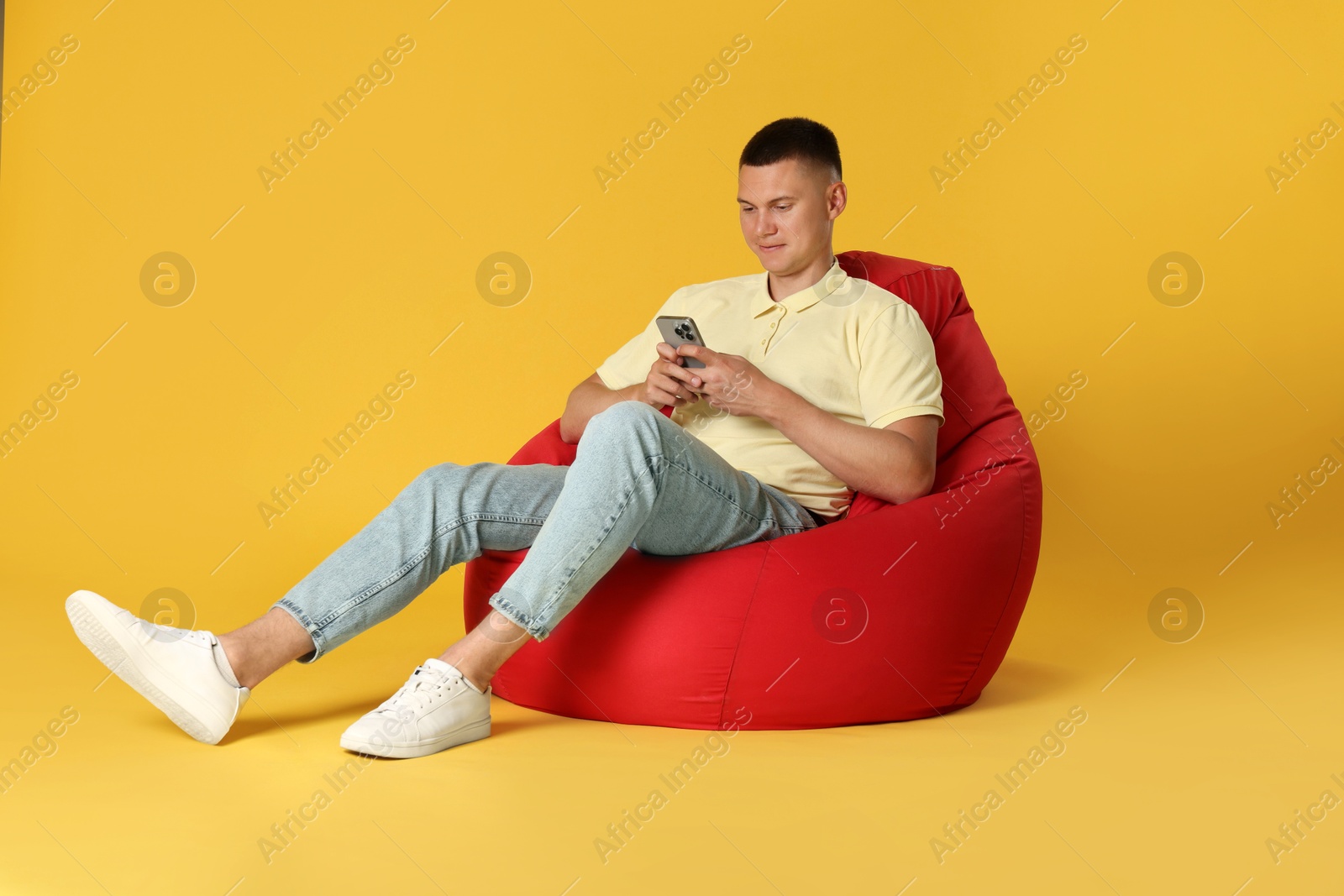 Photo of Handsome man with smartphone on red bean bag chair against yellow background