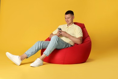 Handsome man with smartphone on red bean bag chair against yellow background