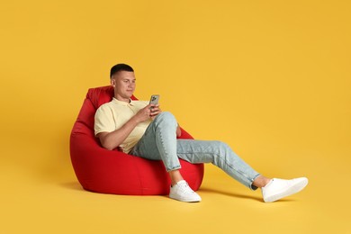 Handsome man with smartphone on red bean bag chair against yellow background