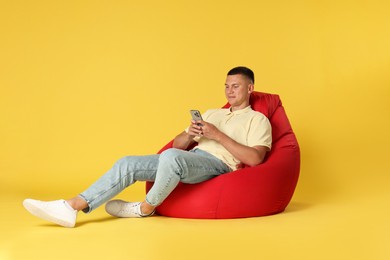 Photo of Handsome man with smartphone on red bean bag chair against yellow background