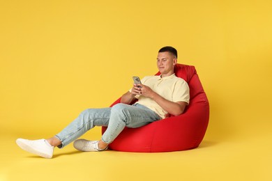 Handsome man with smartphone on red bean bag chair against yellow background