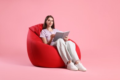 Photo of Beautiful young woman with book sitting on red bean bag chair against pink background