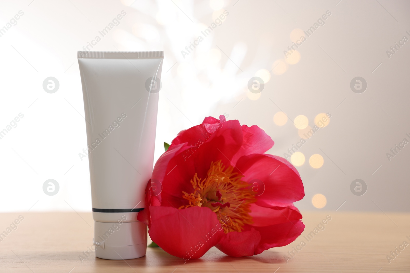 Photo of Tube of cosmetic product and peony flower on wooden table against light background with blurred lights