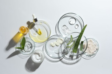 Photo of Petri dishes with different cosmetic products and aloe vera leaves on white background, flat lay