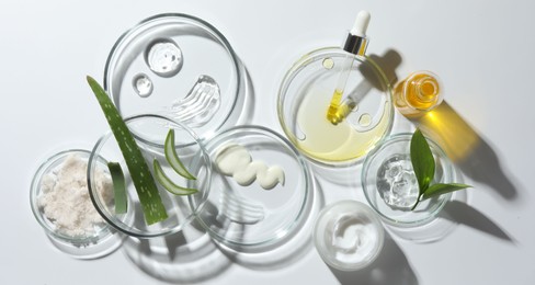 Petri dishes with different cosmetic products and aloe vera leaves on white background, flat lay