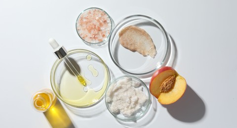 Petri dishes with different cosmetic products and peach on white background, flat lay