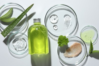 Photo of Petri dishes with different cosmetic products and aloe vera leaves on white background, flat lay