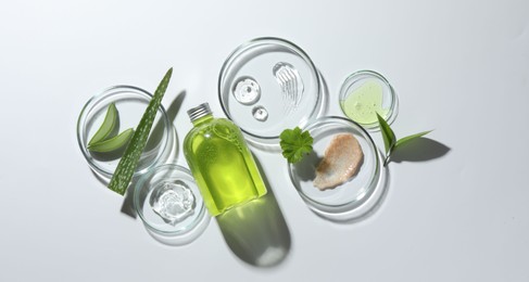 Photo of Petri dishes with different cosmetic products and aloe vera leaves on white background, flat lay