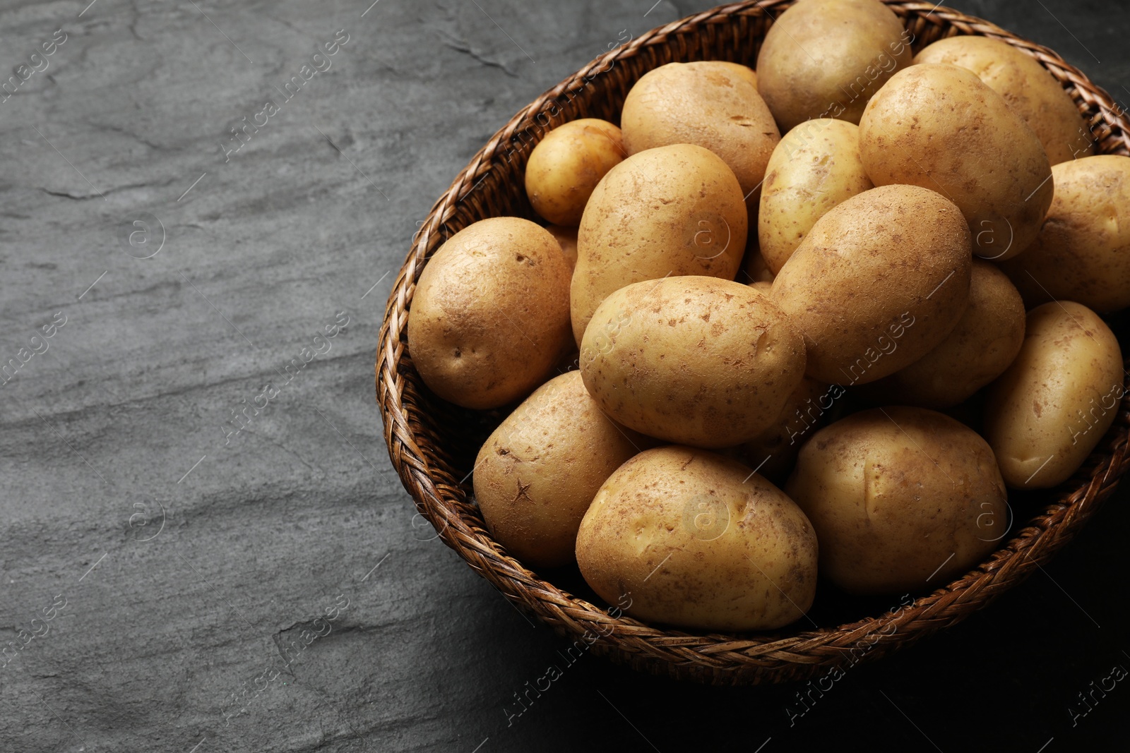 Photo of Many fresh potatoes in wicker basket on black textured table, closeup. Space for text