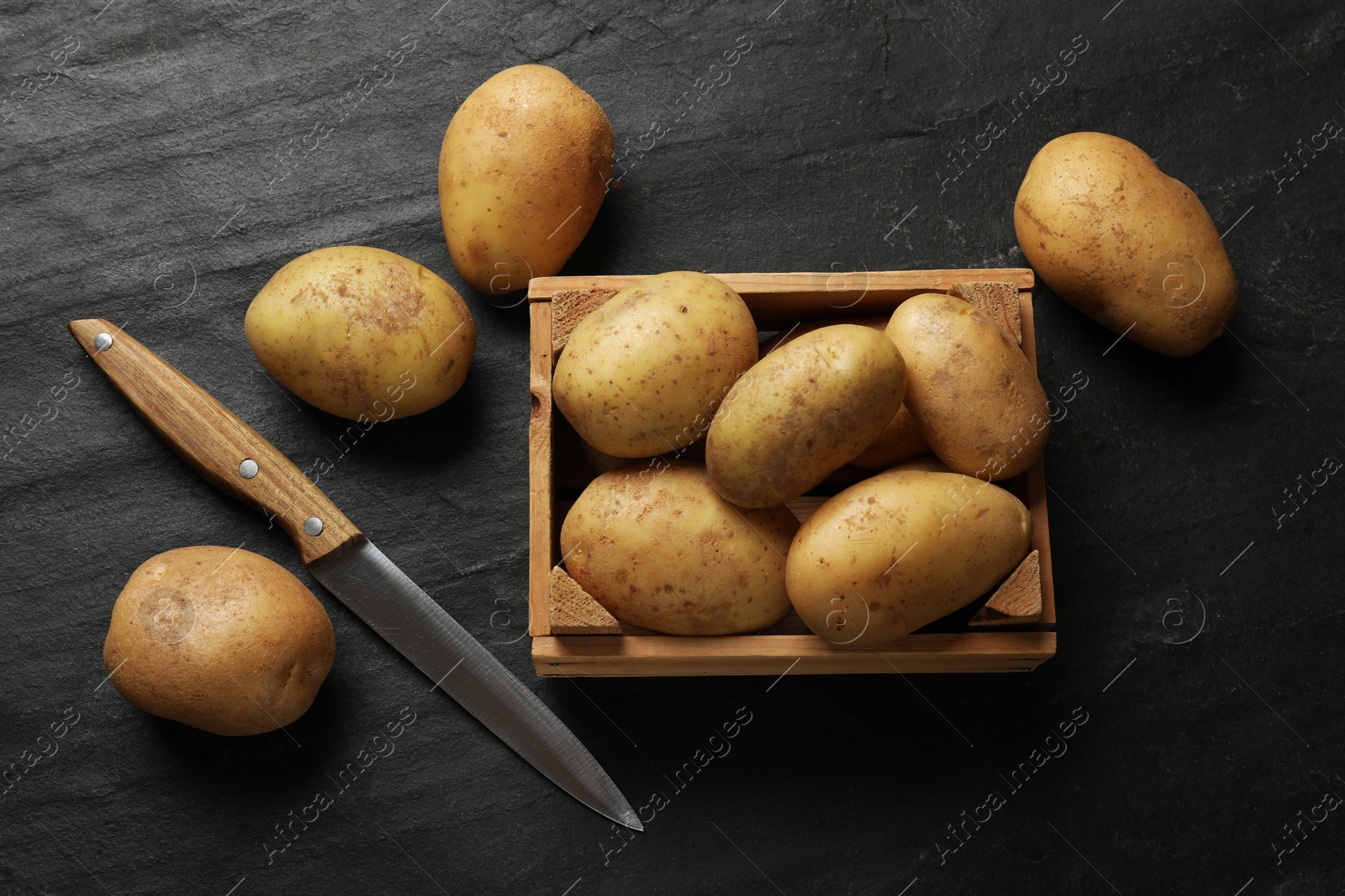 Photo of Many fresh potatoes, wooden crate and knife on black textured table, flat lay