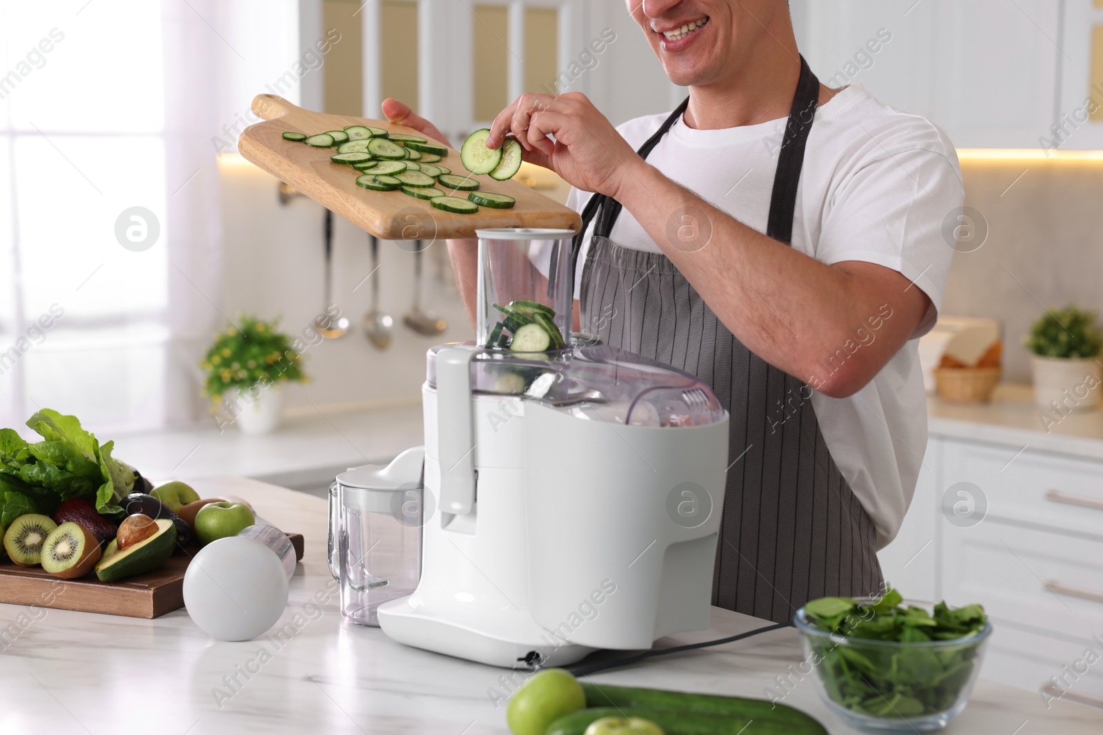 Photo of Man putting fresh cucumber into juicer at white marble table in kitchen, closeup