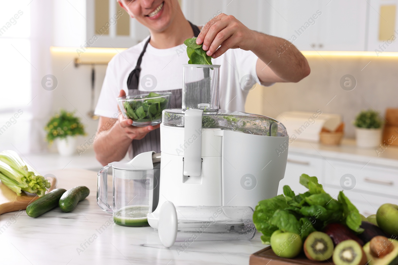 Photo of Man putting fresh basil into juicer at white marble table in kitchen, closeup