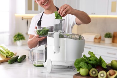 Photo of Man putting fresh basil into juicer at white marble table in kitchen, closeup