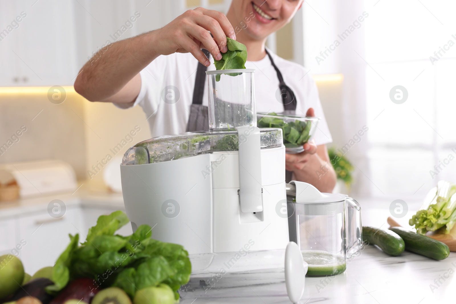 Photo of Man putting fresh basil into juicer at white marble table in kitchen, closeup