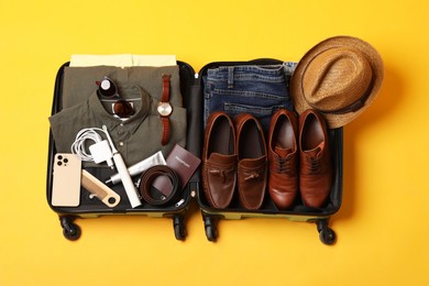 Photo of Open suitcase with traveler's belongings on yellow background, top view