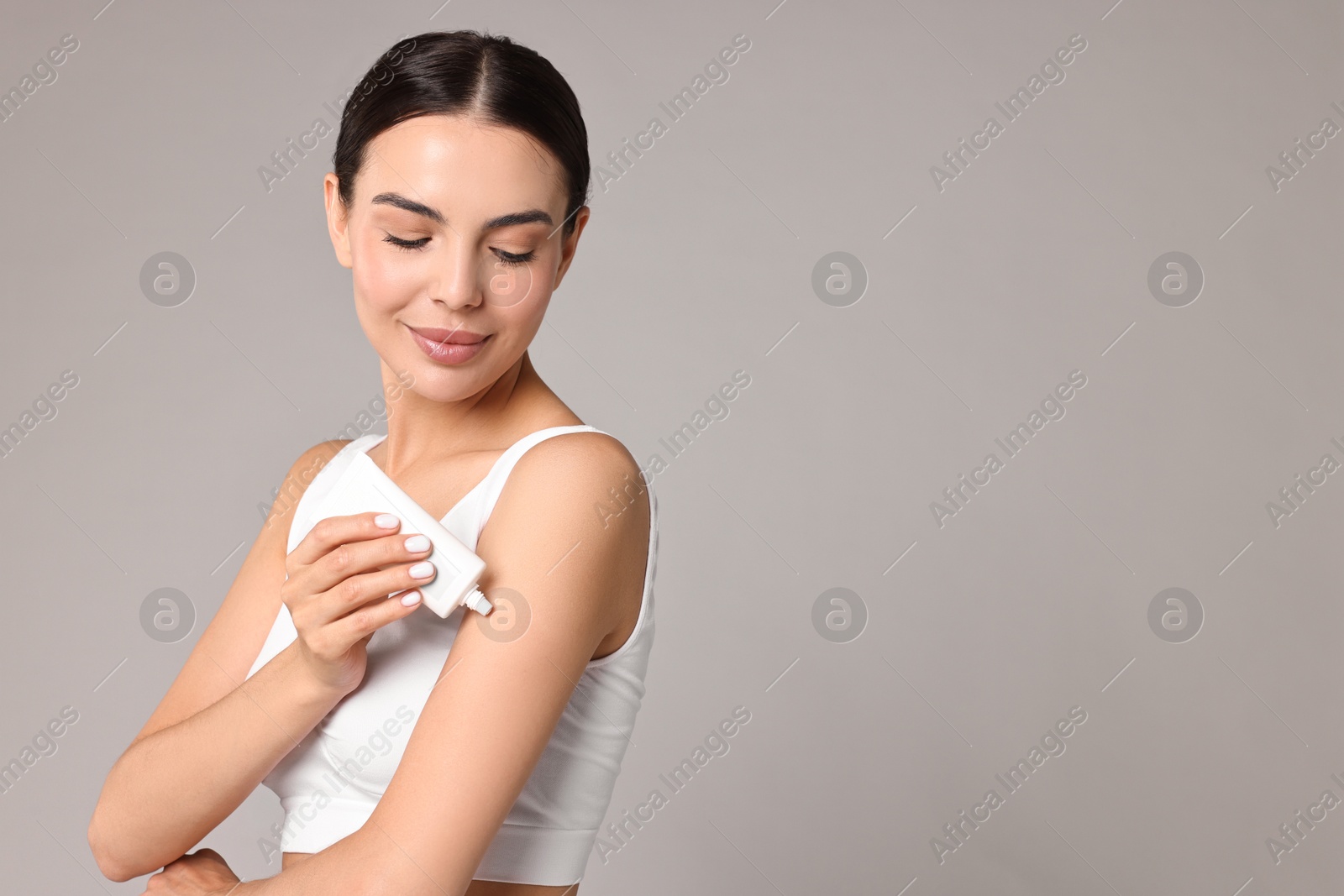 Photo of Beautiful woman applying cream onto arm on grey background, space for text. Body care