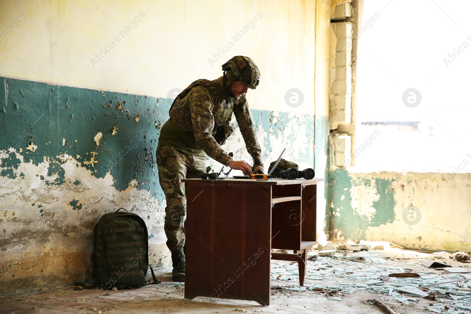 Photo of Military mission. Soldier in uniform using laptop at table inside abandoned building