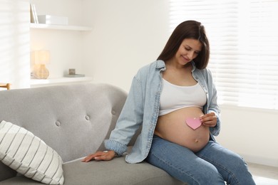 Beautiful pregnant woman with pink paper heart on sofa at home