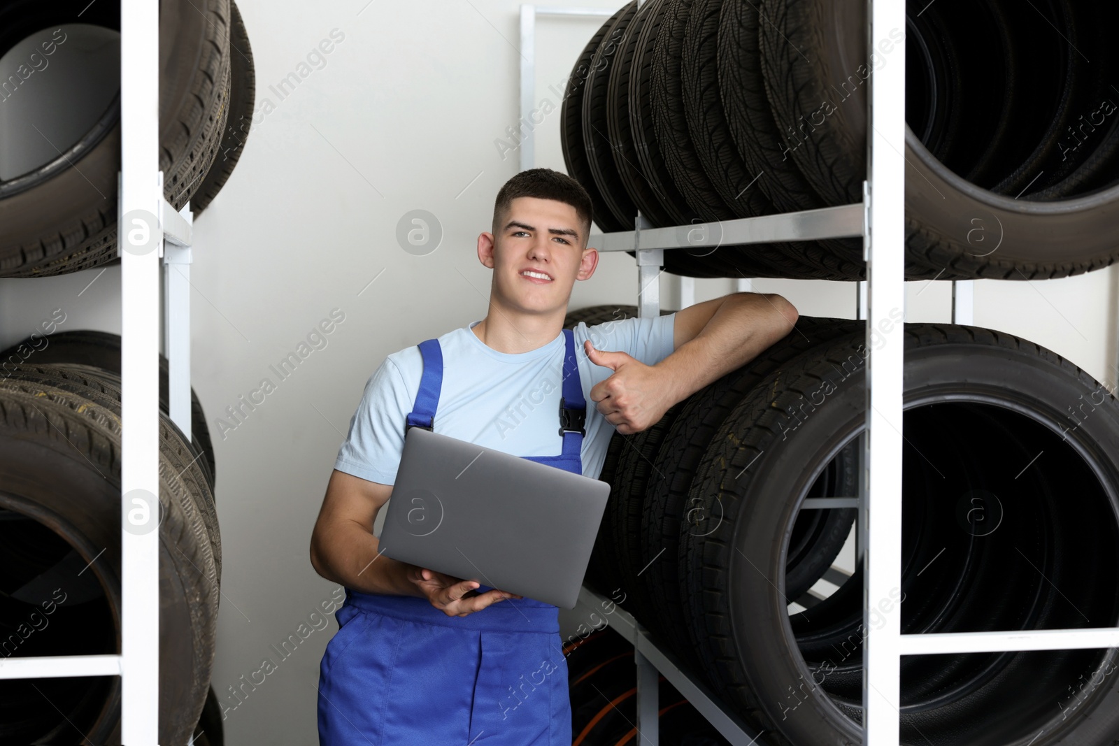 Photo of Smiling young man with laptop near car tires showing thumbs up in auto store