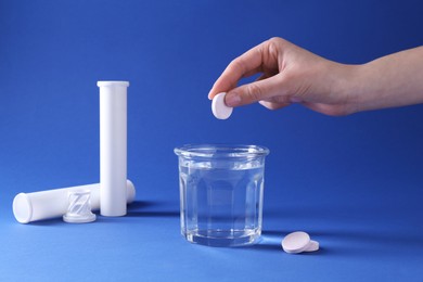 Woman putting effervescent pill into glass of water on blue background, closeup