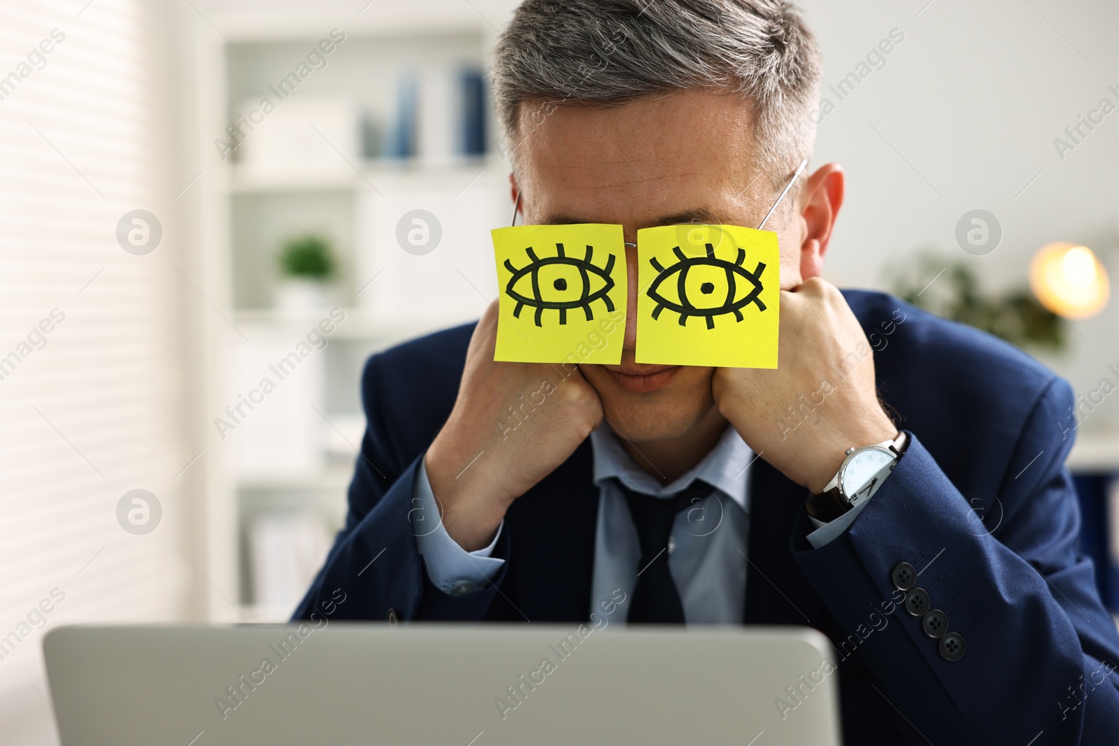 Photo of Man with fake eyes painted on sticky notes snoozing at workplace in office