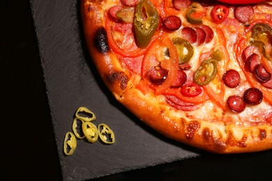 Photo of Delicious pizza Diablo and peppers on slate board against black background, top view