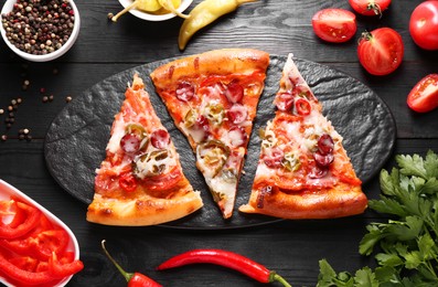 Photo of Slate board with pieces of delicious pizza Diablo among products on black wooden table, flat lay