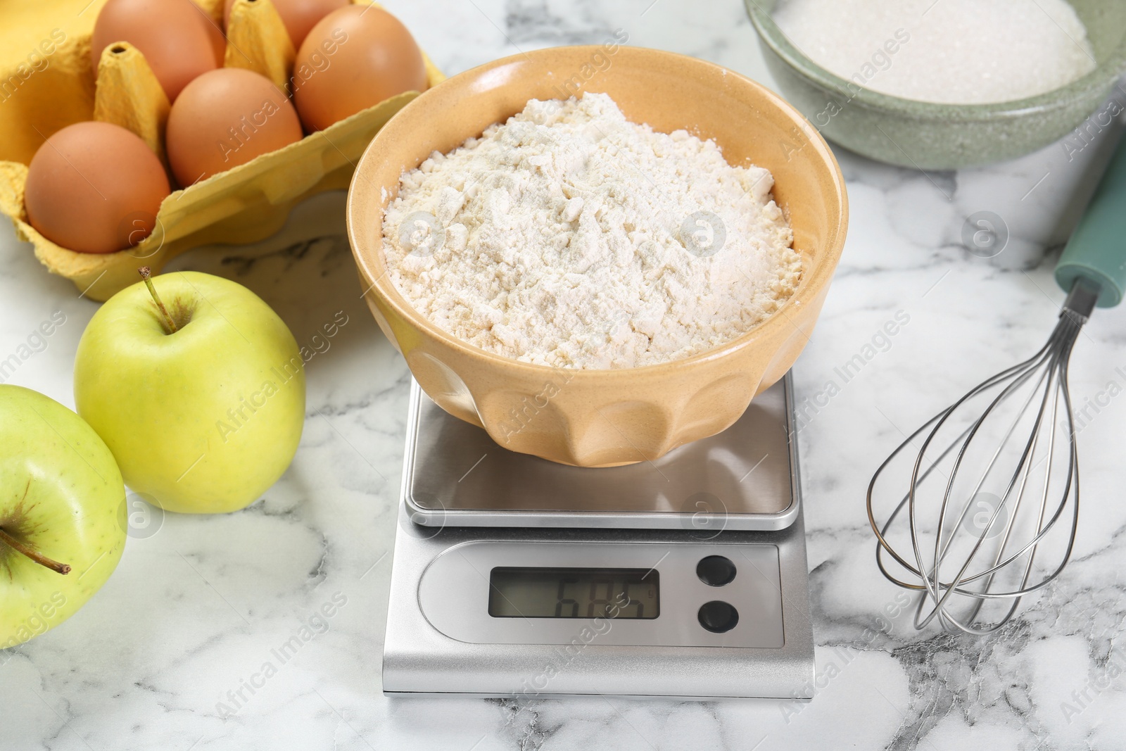 Photo of Kitchen scale with bowl of flour, whisk and products on white marble table, closeup