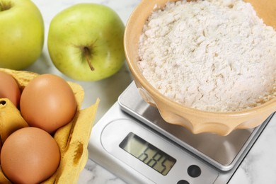 Photo of Kitchen scale with bowl of flour, eggs and apples on white table, closeup